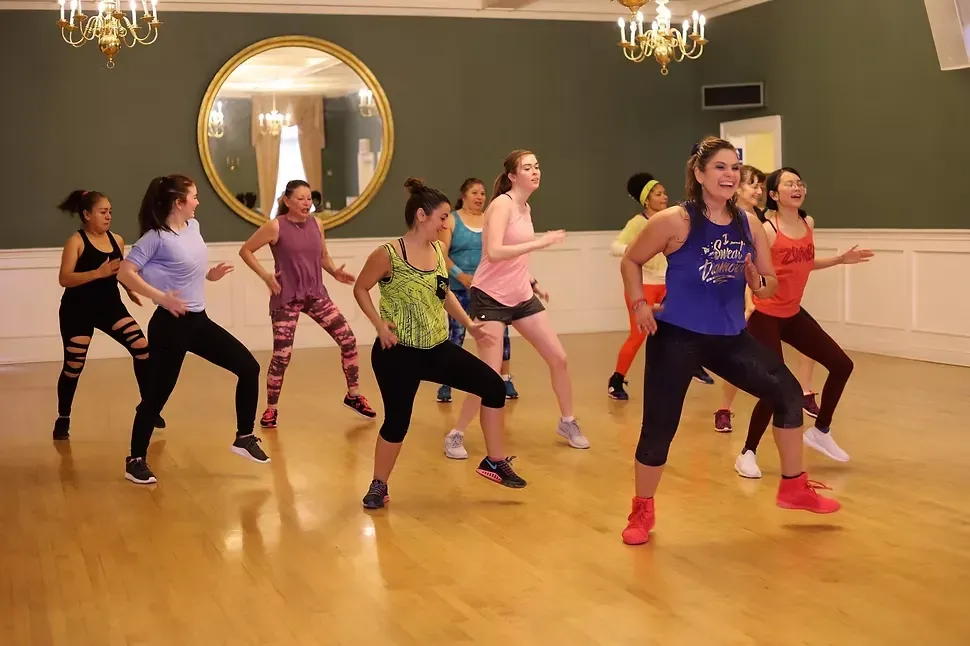 A Zumba with Ida class dancing together in a hall with chandeliers and a wooden floor